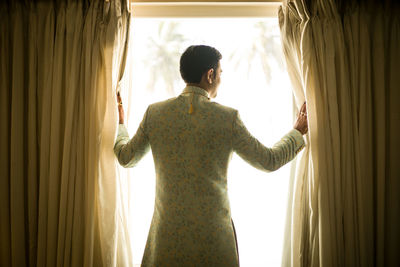 Rear view of man looking through window at home