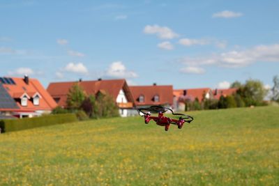 Quadcopter flying over grassy field against houses and sky