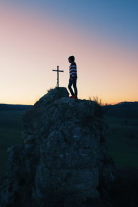 Boy standing by cross on mountain against sky during sunset