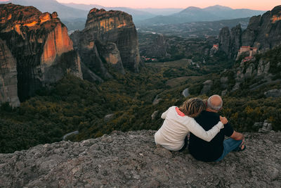 Rear view of couple sitting on cliff against mountains