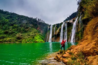 Rear view of man standing by waterfall against sky