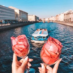 Cropped hands of person holding wineglasses with roses against river in city