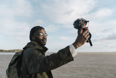 Man making vlog of himself at beach against sky