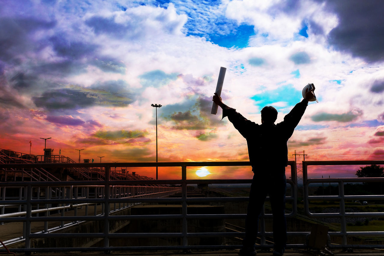 SILHOUETTE MAN STANDING BY RAILING AGAINST ORANGE SKY