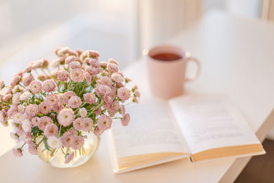 Small pink flowers bouquet in vase with  background of pink cup of tea or coffee and opened book