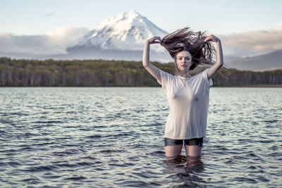 Portrait of young woman tossing hair while standing in lake against sky during sunset