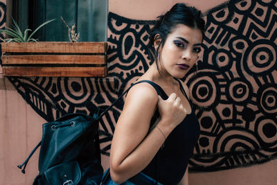 Portrait of young woman with backpack standing against patterned wall