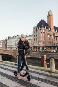 Full length of teenage girl talking on mobile phone while standing on e-scooter by canal in city