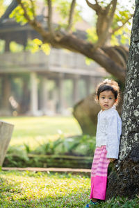 Portrait of girl standing on grass by tree trunk against building