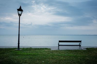 Bench on street light by sea against sky
