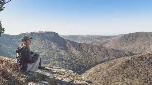 Thoughtful woman looking at mountains while sitting on rock against clear sky