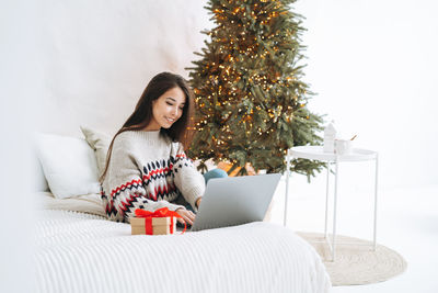 Young asian woman with dark long hair using laptop on bed in room with christmas tree at home