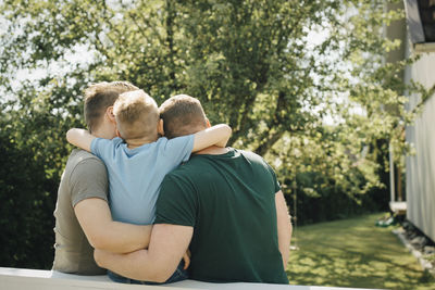 Rear view of son with arm around fathers sitting on fence in yard