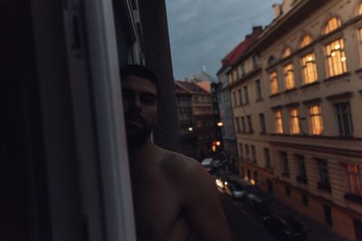 Portrait of shirtless man standing at window in city