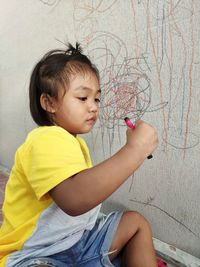 Girl holding crayon while sitting by wall