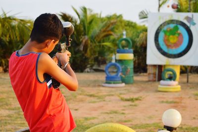 Rear view of boy aiming paintball gun on target against trees