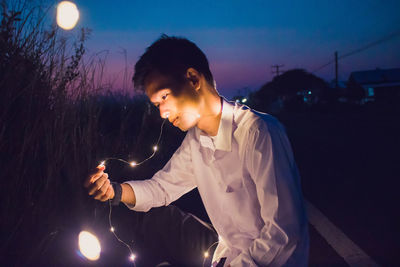 Young man holding illuminated string light while sitting on field during dusk