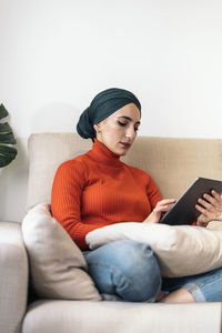 Muslim female in casual clothes and headscarf sitting cross legged on sofa and browsing social media on tablet while spending time in light living room at home