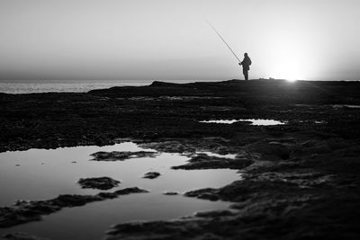 Silhouette fisherman fishing at sea shore against sky during sunset