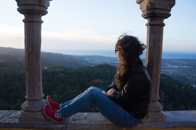 Side view of woman sitting on retaining wall against sky