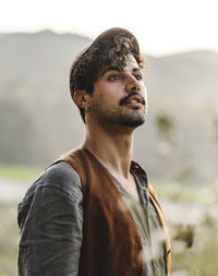 Dreamy young curly haired bearded ethnic male with piercing in ear wearing stylish clothes and cap looking away while standing on green meadow against blurred mountains