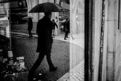 Man with umbrella walking on footpath reflecting in glass window