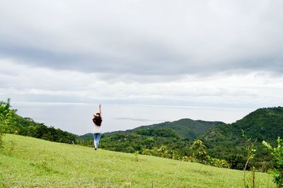 Rear view of woman showing peace sign while standing on grassy field against cloudy sky