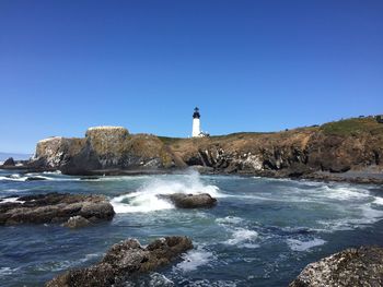 Lighthouse on rocks by sea against clear blue sky