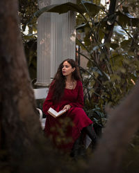 Portrait of young woman with a book in the long red dress sitting in the nature