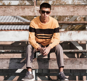 Asian teenage boy in yellow t-shirt and wear black glasses, sit on wooden stage outdoors. 