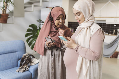 Pregnant woman sharing smart phone with friend in living room