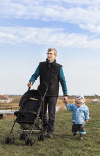 Man holding hands with granddaughter while walking on grassy field against sky