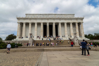 People walking in front of historical building against cloudy sky