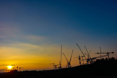 Silhouette cranes by building against sky during sunset