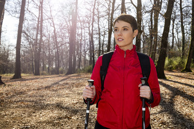 Young woman has short hair, wears a red jacket and uses trekking poles for a hike in the woods