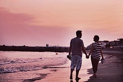 Rear view of men walking on beach against sky during sunset