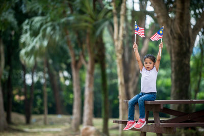 Portrait of girl holding malaysian flag while sitting on bench at park