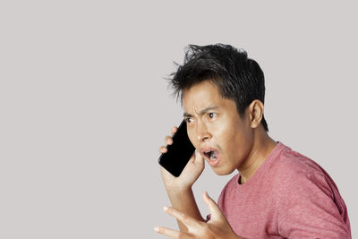 Young man talking on the phone, shocked with and surprise face on gray background.