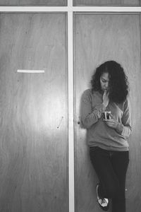 Thoughtful woman with finger on lips using phone while leaning on wooden door