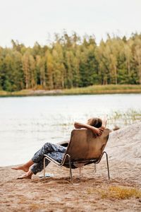 Young woman relaxing on chair at lakeshore against sky