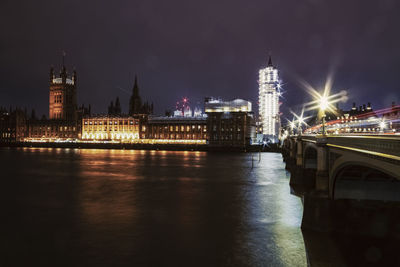 Illuminated bridge over river by buildings against sky at night