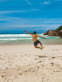 Rear view of shirtless young man jumping in air, clicking heels on tropical sandy beach