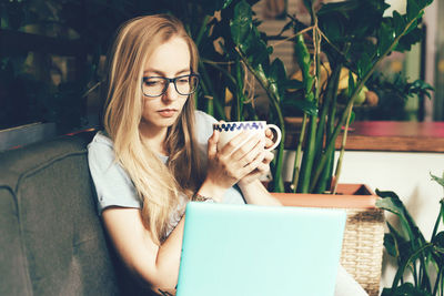 A student girl in glasses is holding a coffee mug and surfing the internet in a laptop.