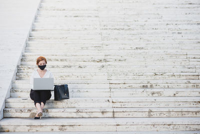 Businesswoman in formal wear and protective mask sitting on stone steps in city and working on netbook during covid 19 epidemic