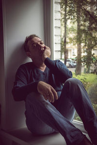 Portrait of sad mature man with hand on chin sitting at window sill