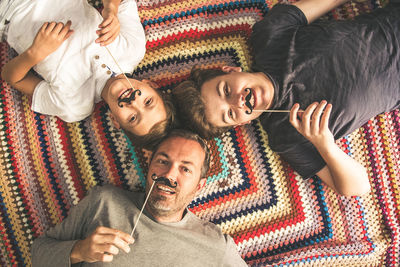 Portrait of father with sons holding prop mustache while lying on floor