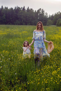 Mother and daughter in dresses and a hat stand in a field of yellow flowers in the summer day