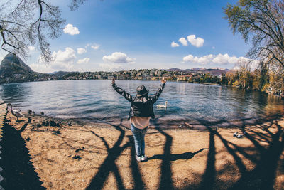 Rear view of cheerful woman standing at lakeshore against sky