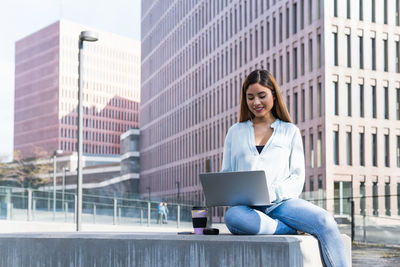 Smiling businesswoman using laptop sitting against building