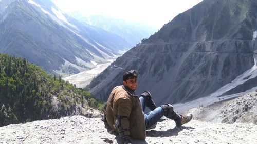 Portrait of happy young man sitting on cliff against mountains